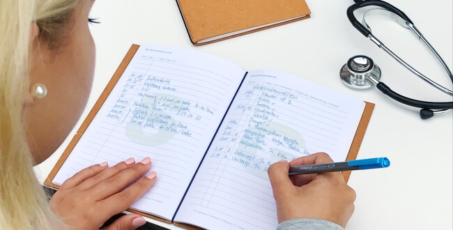Eine Person dokumentiert handschriftlich ihre Gesundheitshistorie in einem aufgeschlagenen Notizbuch. Neben dem Heft liegen ein geschlossener Planer und ein Stethoskop auf dem weißen Schreibtisch, was den medizinischen Kontext der Dokumentation unterstreicht.