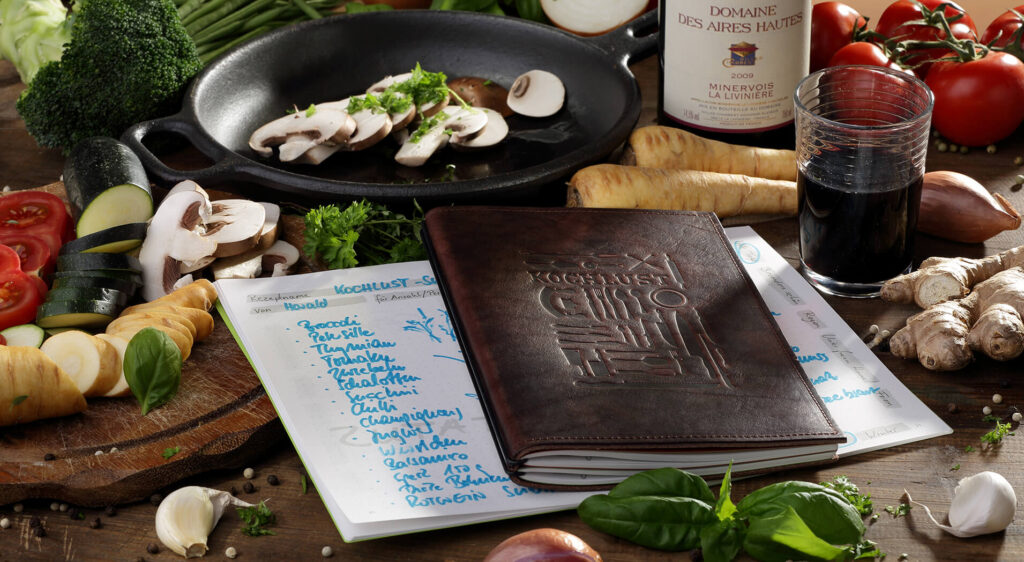 Ein hochwertiges X17 Rezeptbuch aus braunem Leder im A5 Hochformat, liegend auf einem Küchentisch inmitten von frischen Zutaten wie Brokkoli, Tomaten, Champignons und einer Flasche Rotwein.