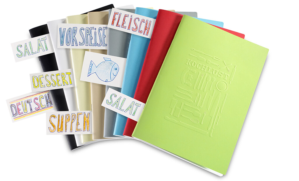 Mehrere farbenfrohe Hefte des Kochlust Rezeptbuch-Systems fächerförmig angeordnet, mit handgezeichneten Registern für Kategorien wie Fleisch, Vorspeise, Salat, Dessert, Deutsch und Suppen.