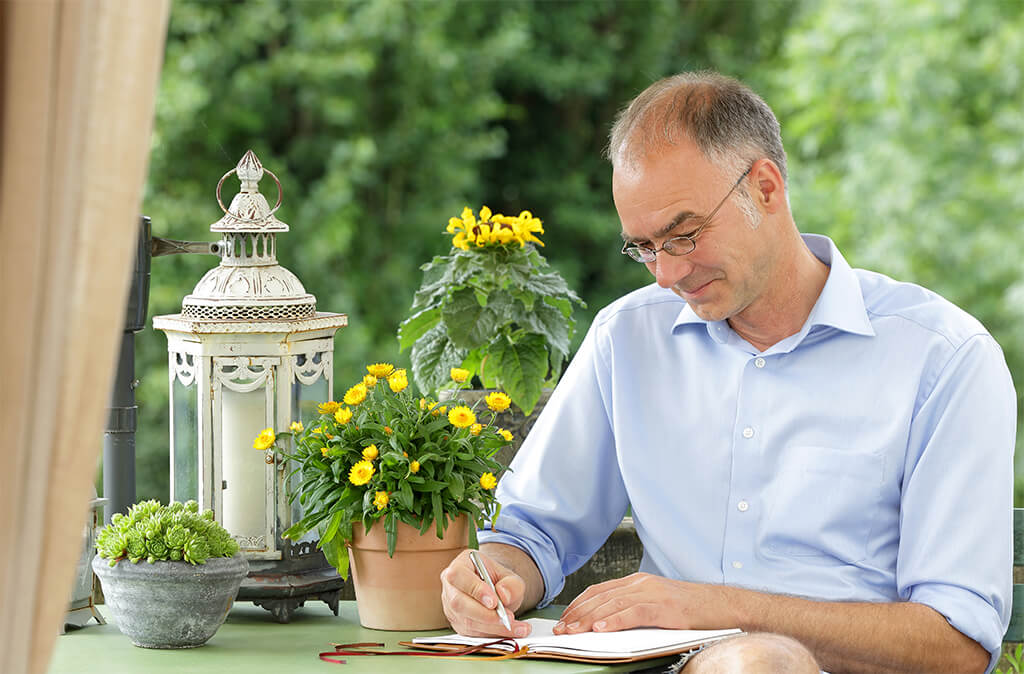 Ein konzentrierter Mann schreibt in ein Notizbuch an einem Gartentisch – ein Beispiel für Selbstführung und die Etablierung täglicher Routinen und Tracking. Die ruhige Umgebung mit Blumen und einer Laterne unterstreicht ein fokussiertes Mindset und die Bedeutung von Struktur im (Arbeits-)Alltag.