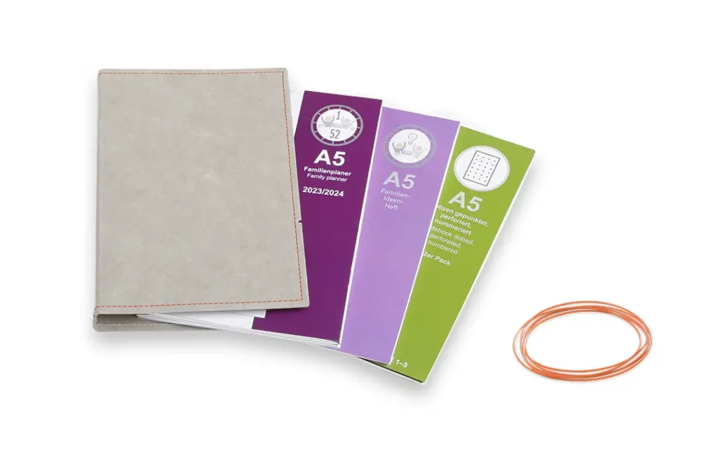 Ein vierteiliges X17 Set eines A5-Familienplaners bestehend aus einer grauen Hülle mit orangefarbener Naht, drei verschiedenen Einlegeheften in Lila und Grün sowie orangefarbenen Verschlussbändern.