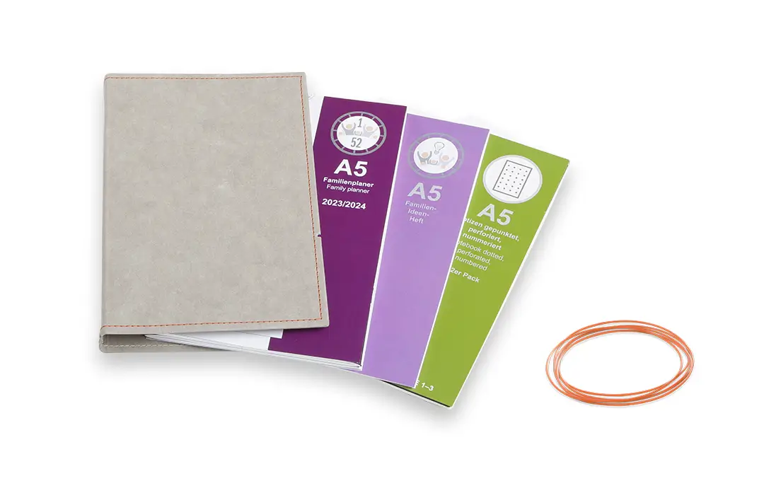 Ein vierteiliges X17 Set eines A5-Familienplaners bestehend aus einer grauen Hülle mit orangefarbener Naht, drei verschiedenen Einlegeheften in Lila und Grün sowie orangefarbenen Verschlussbändern.