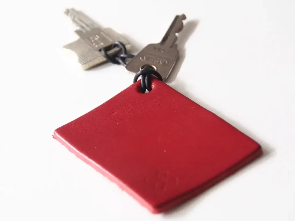 Nahaufnahme eines Schlüsselanhägers mit einem quadratischen, roten Lederanhänger. An dem Anhänger sind über schwarze Ringe zwei silberne Metallschlüssel befestigt.