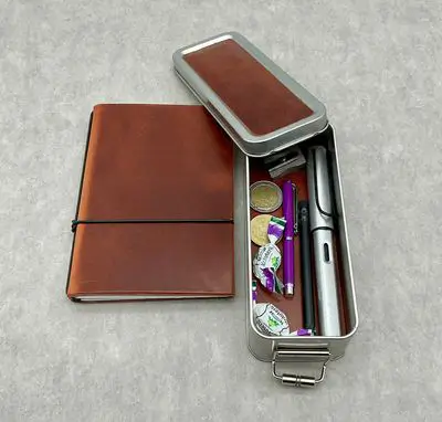 Draufsicht auf die X17 Sonderedition Box aus Metall mit passendem Leder-Notizbuch. Die geöffnete Stiftebox ist mit Schreibutensilien wie einem Lamy-Füller, Stiften, einem Anspitzer und kleinen Süßigkeiten gefüllt.