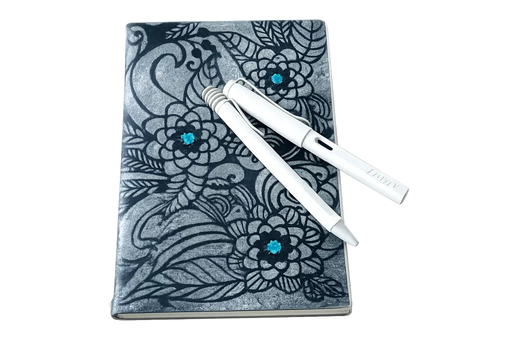 Ein X17 Notizbuch aus hellem Texon. Das Cover ist mit schwarzen floralen Ornamenten und Blüten verziert, deren Zentren durch türkisfarbene Akzente hervorgehoben werden. Darauf liegen zwei weiße Lamy-Schreibgeräte diagonal übereinander.